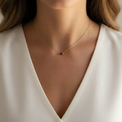 A close-up of a model wearing a delicate gold chain necklace with a small rectangular purple gemstone pendant, paired with a white V-neck top.