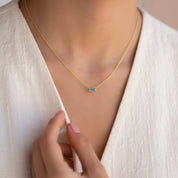 A close-up lifestyle shot of a delicate gold chain necklace with a small rectangular green blue gemstone pendant, worn by a model in a white linen V-neck top.
