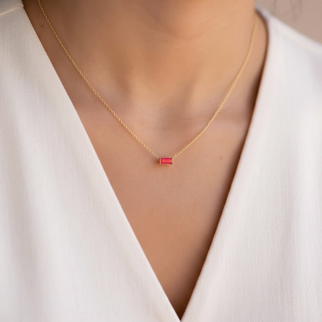 A close-up of a model wearing a delicate gold chain necklace with a small rectangular red gemstone pendant, styled with a white V-neck top.