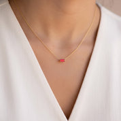 A close-up of a model wearing a delicate gold chain necklace with a small rectangular red gemstone pendant, styled with a white V-neck top.