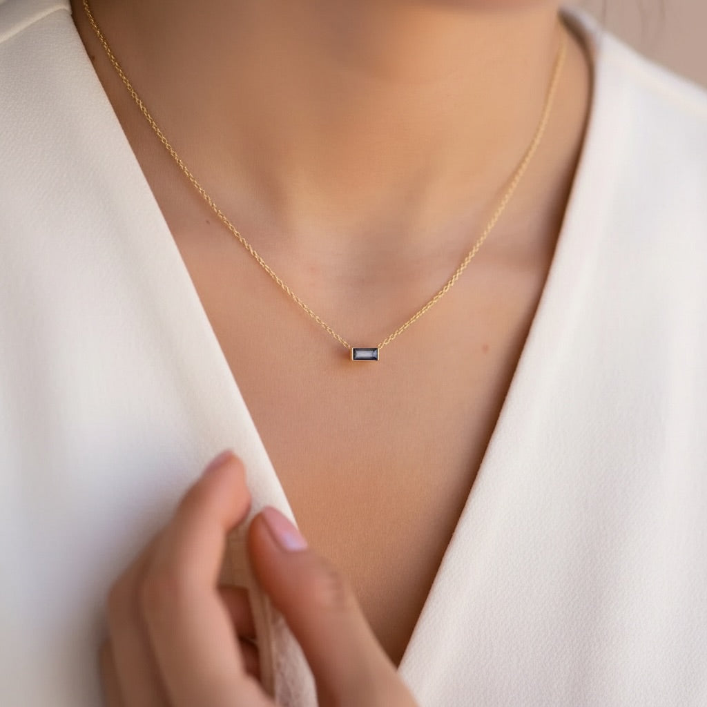A close-up of a delicate gold chain necklace with a small rectangular dark blue gemstone pendant, worn by a model in a white V-neck top.
