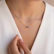 A close-up of a delicate gold chain necklace with a small rectangular dark blue gemstone pendant, worn by a model in a white V-neck top.