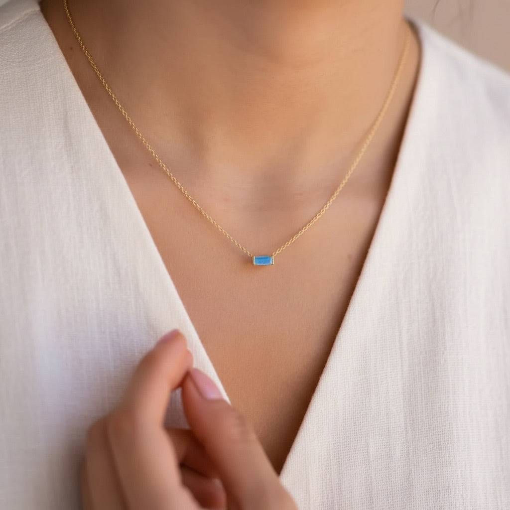 A close-up of a delicate gold chain necklace with a small rectangular blue stone pendant, worn by a model in a white V-neck top.