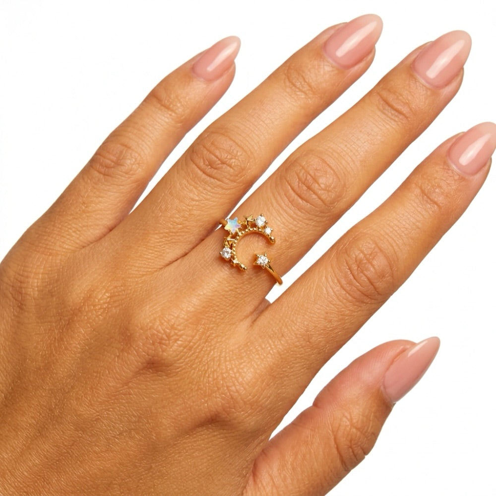 A gold crescent moon ring with small clear gemstones, shown worn on a hand with manicured nails against a white background.