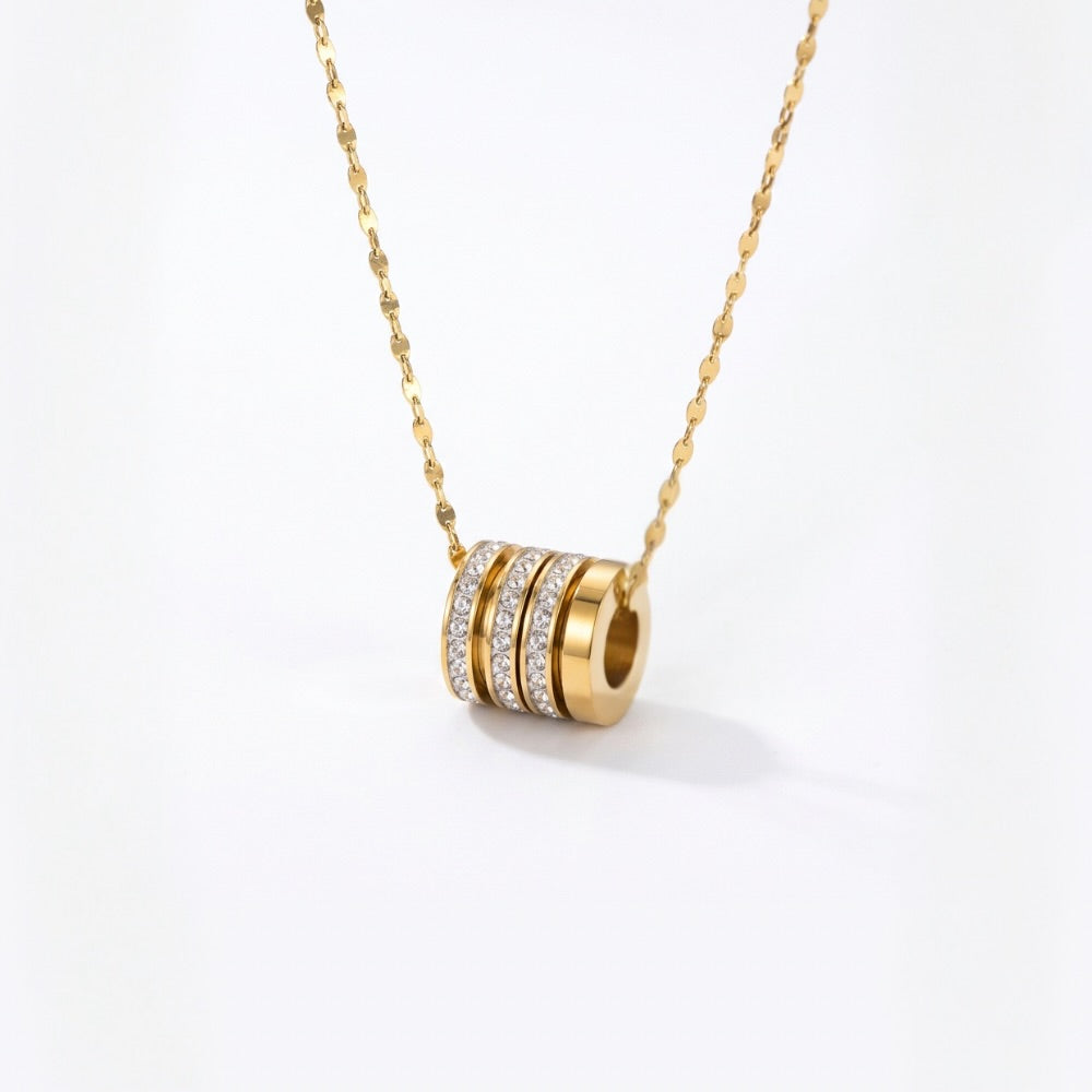 A gold chain necklace with a cylindrical pendant featuring three horizontal rows of small clear stones on a gold-toned base, photographed on a clean white background.