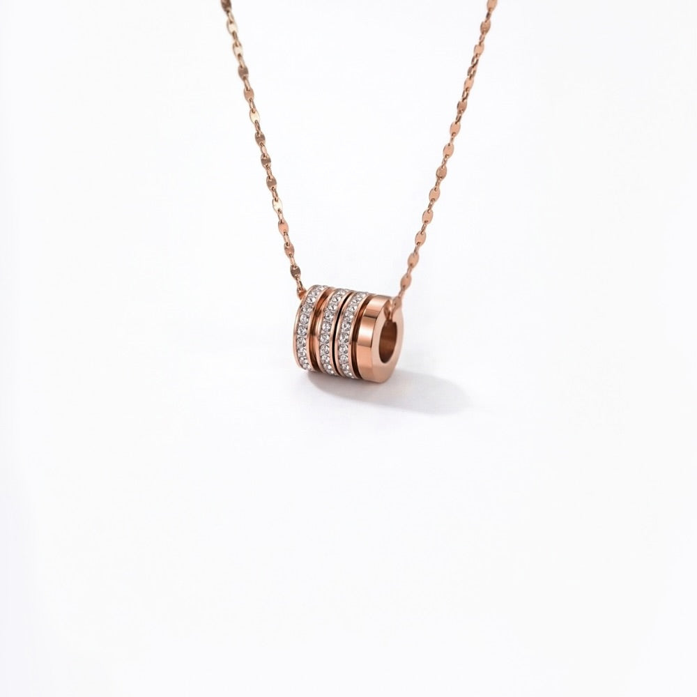 A rose gold necklace with a cylindrical pendant featuring three stacked bands, with the center band set with small clear stones, shown against a plain white background.