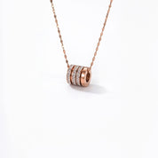 A rose gold necklace with a cylindrical pendant featuring three stacked bands, with the center band set with small clear stones, shown against a plain white background.