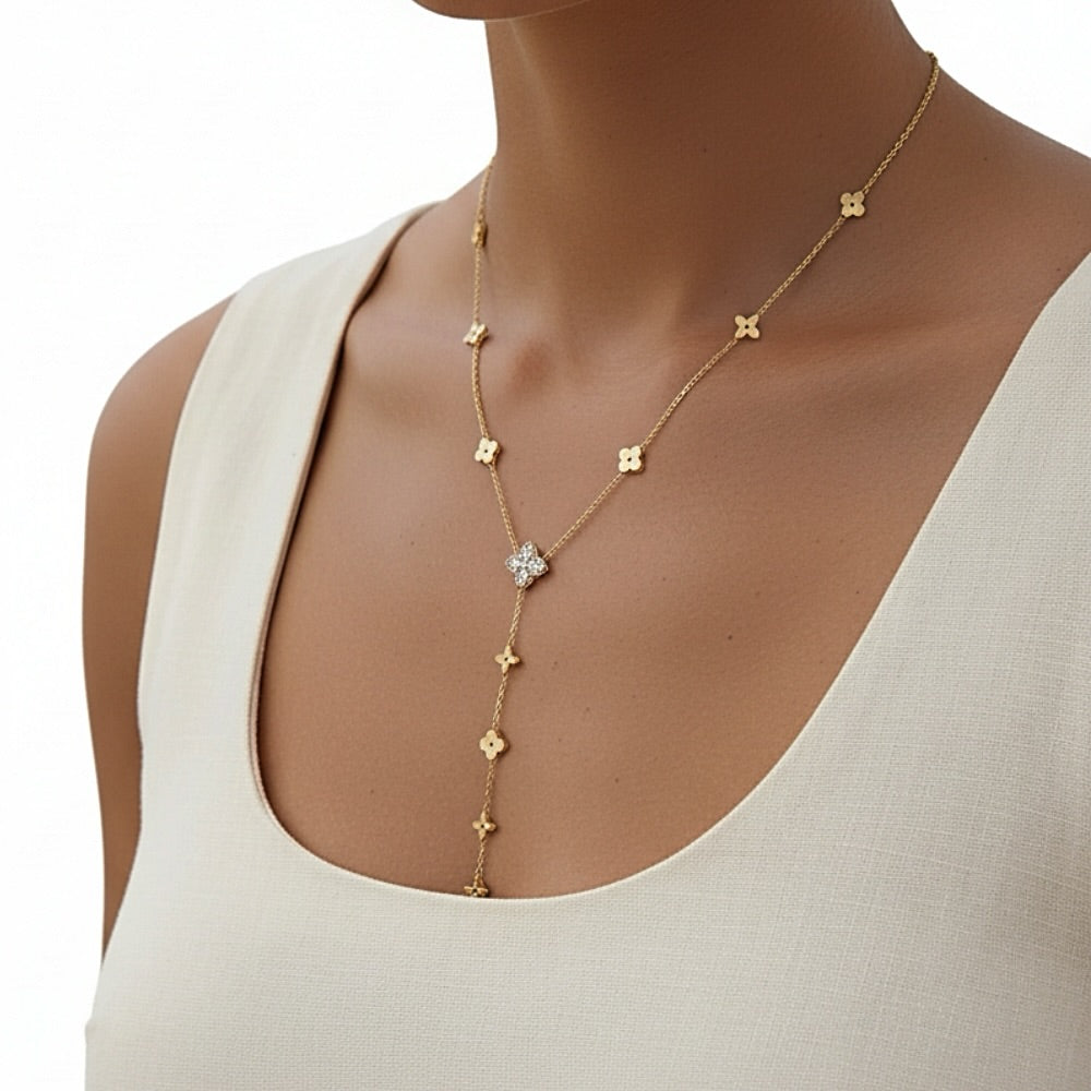 A gold Y-necklace with small clover motifs and a central clear crystal flower, shown on a model wearing a cream-colored top against a white background.
