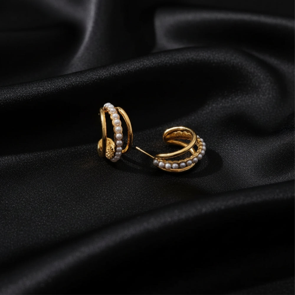 A pair of gold huggie hoop earrings adorned with rows of white faux pearls, displayed on a draped black satin fabric background.