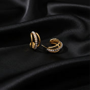 A pair of gold huggie hoop earrings adorned with rows of white faux pearls, displayed on a draped black satin fabric background.