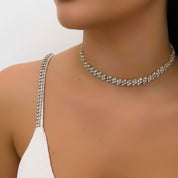 A close-up of a model wearing a gold rhinestone choker necklace and a white spaghetti-strap top against a plain light background.