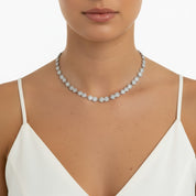 A close-up of a model wearing a delicate silver tennis necklace adorned with small round-cut diamonds, styled with a white camisole top against a clean white background.