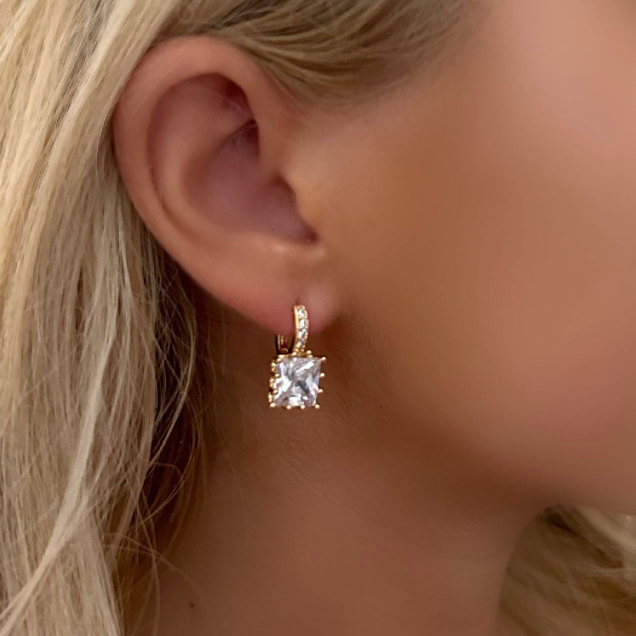 A close-up of a model's ear wearing a small gold huggie hoop earring with a clear square-cut gemstone.