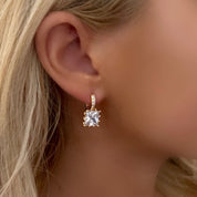 A close-up of a model's ear wearing a small gold huggie hoop earring with a clear square-cut gemstone.