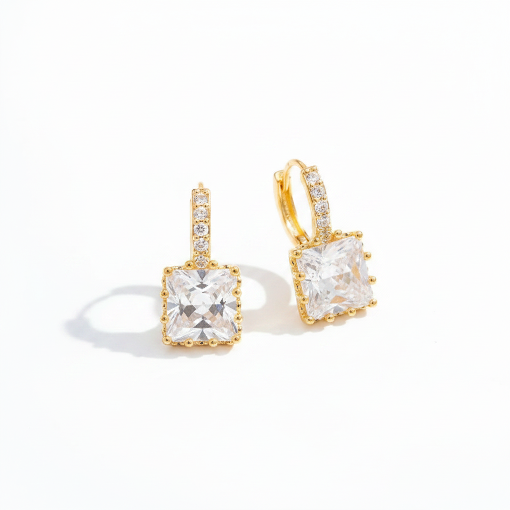 A pair of gold huggie hoop earrings with a square princess-cut clear gemstone in a gold bezel setting, accented with small pavé-set clear stones on the upper band, shown on a white background.