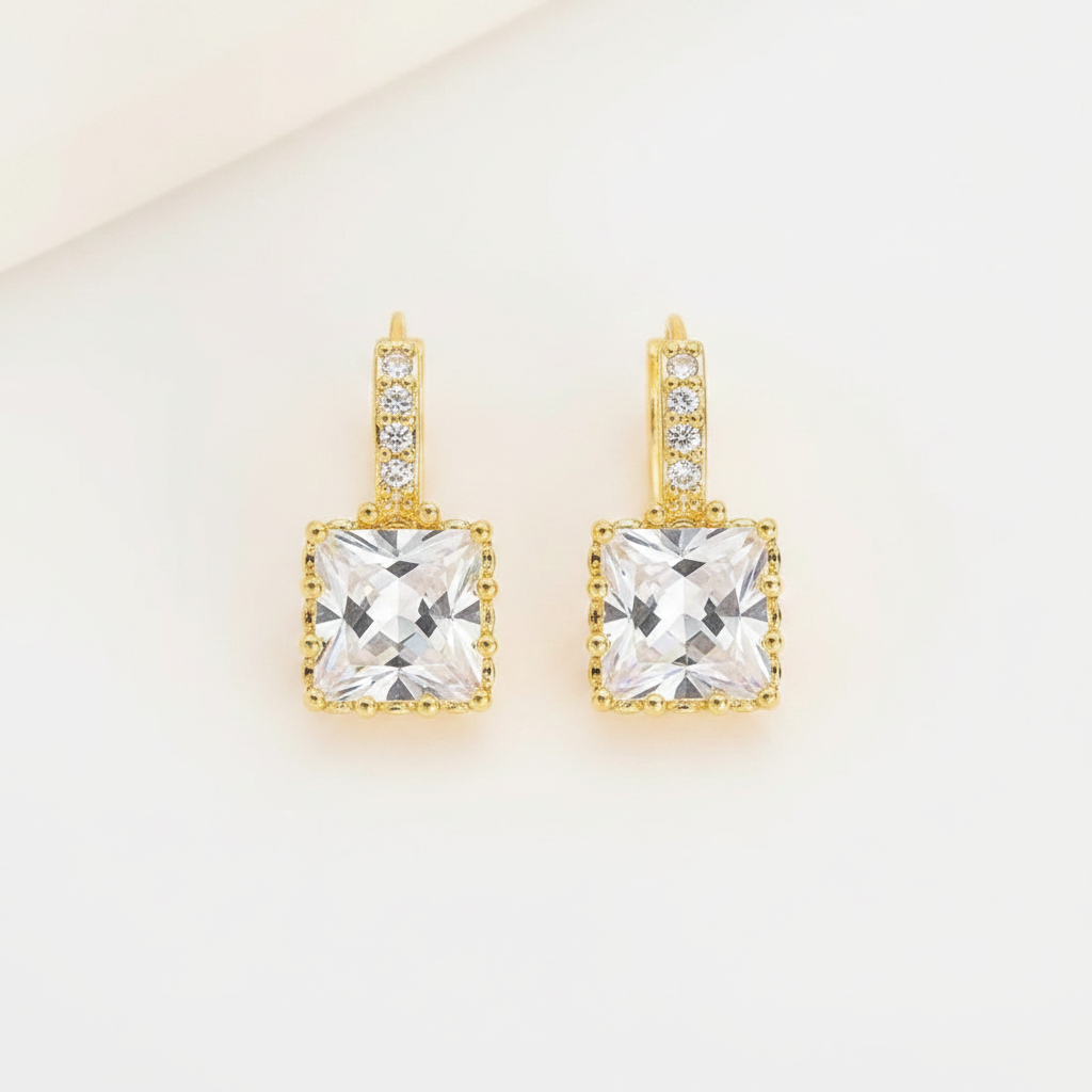 A pair of gold drop earrings featuring a large square clear crystal pendant with a beaded border, accented by small clear stones along the top bar, set against a clean off-white background.