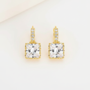 A pair of gold drop earrings featuring a large square clear crystal pendant with a beaded border, accented by small clear stones along the top bar, set against a clean off-white background.