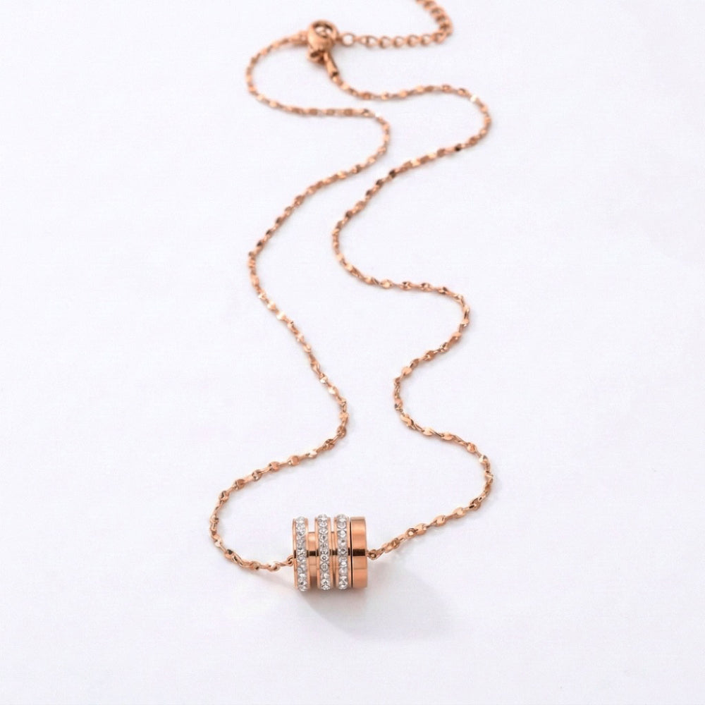 A rose gold chain necklace with a cylindrical pendant featuring three horizontal rows of small clear stones, displayed on a clean white background.