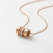 A rose gold necklace with a cylindrical pendant featuring textured bands and a central row of clear pavé stones, displayed on a clean white background.