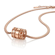 Rose gold snake chain necklace with a cylindrical cutout charm featuring Roman numerals, displayed on a reflective white surface.