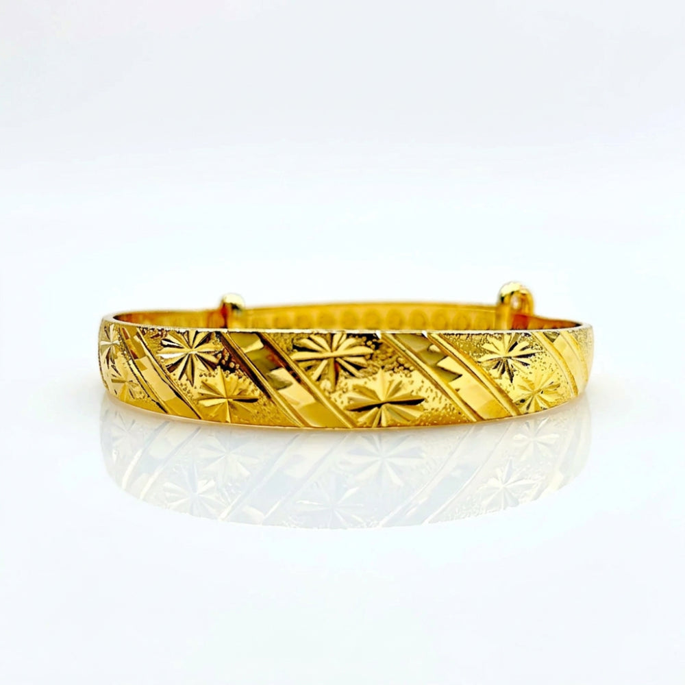 A gold-plated bangle bracelet with a textured, engraved pattern of diagonal stripes and starbursts, displayed on a clean white reflective surface.