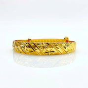 A gold-plated bangle bracelet with a textured, engraved pattern of diagonal stripes and starbursts, displayed on a clean white reflective surface.