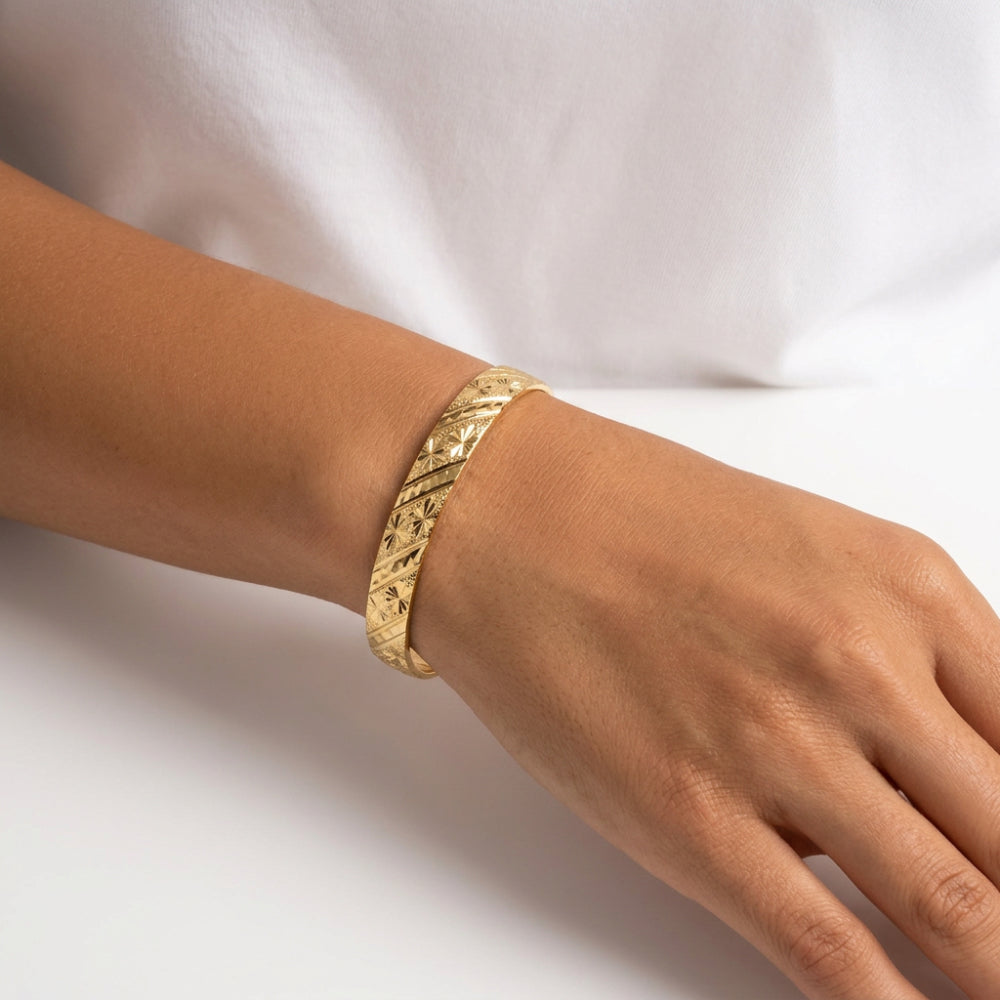 A close-up of a person's wrist wearing a textured gold bangle bracelet against a white background.
