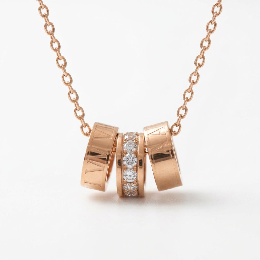 Rose gold necklace with a pendant made of three interlocking cylindrical rings, with the center ring set with clear gemstones, on a white background.