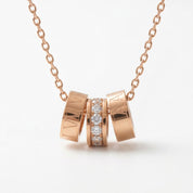 Rose gold necklace with a pendant made of three interlocking cylindrical rings, with the center ring set with clear gemstones, on a white background.