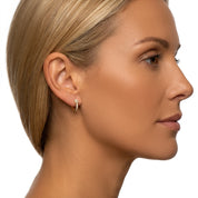 A close-up side profile of a woman with blonde hair wearing a small gold hoop earring with clear stones, set against a plain white background.