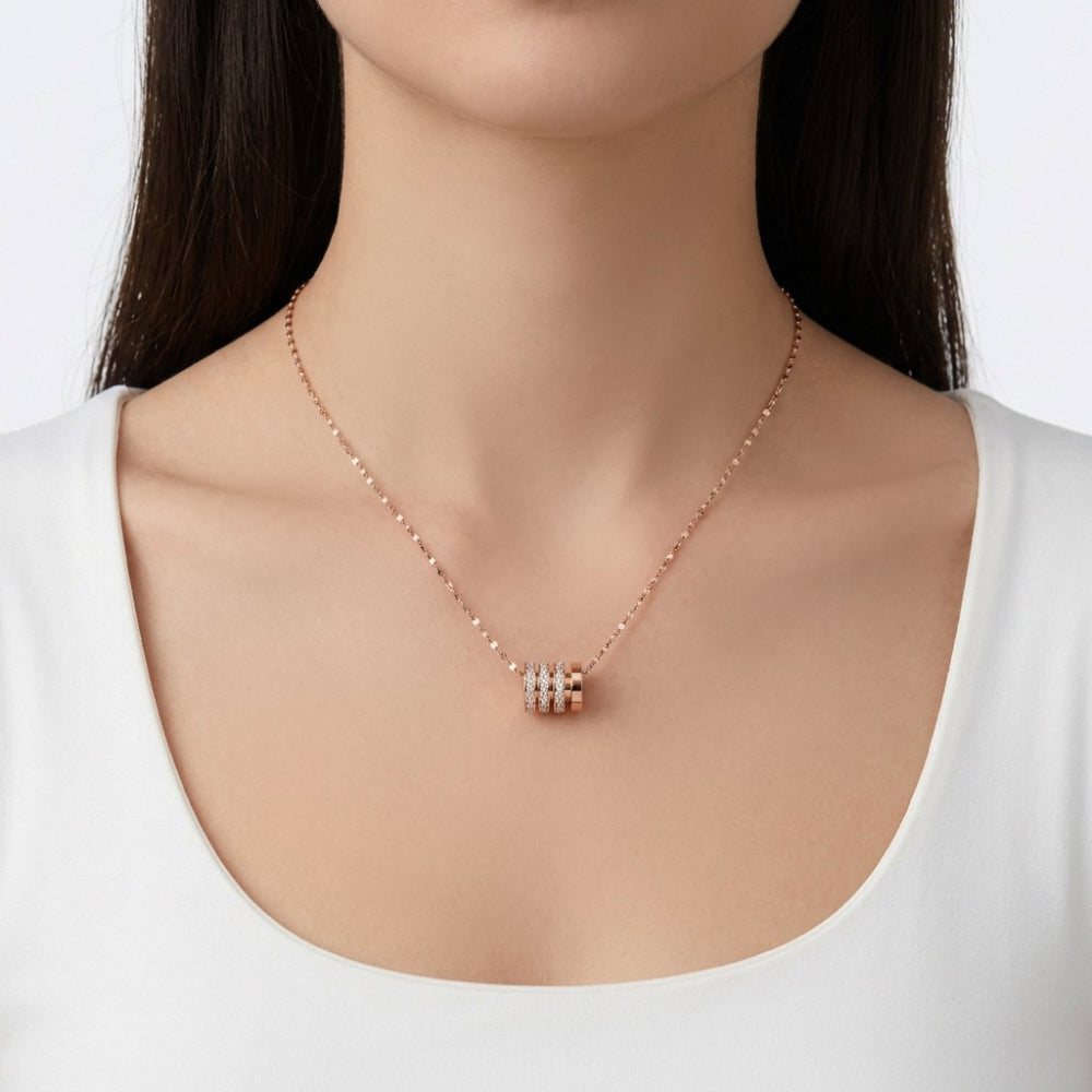 A close-up of a model wearing a delicate rose gold chain necklace with a small cylindrical pendant featuring three horizontal rows of clear stones.