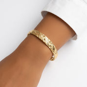 A gold-toned textured bangle bracelet with a carved geometric pattern, shown worn on a person's wrist against a clean white background.