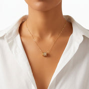 A close-up of a model wearing a delicate gold chain necklace with a small cylindrical pendant featuring three rows of clear stones, styled with a white collared shirt against a clean white background.