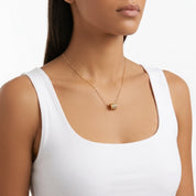 A close-up of a model wearing a delicate gold chain necklace with a small gold and white beaded pendant, styled with a white ribbed tank top against a clean white background.