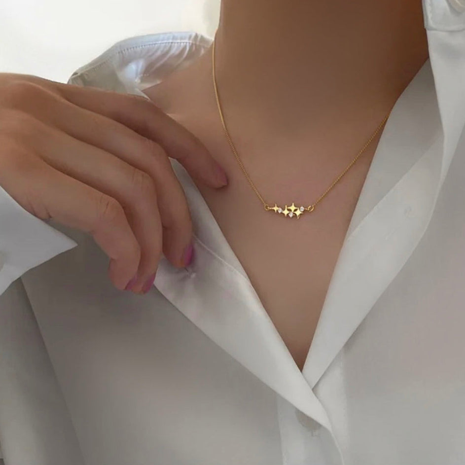 A close-up of a person wearing a delicate gold necklace with a small geometric star-like pendant, styled with a white collared shirt.