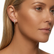 A close-up side profile of a woman with blonde hair wearing a small gold hoop earring with clear stones, set against a plain white background.