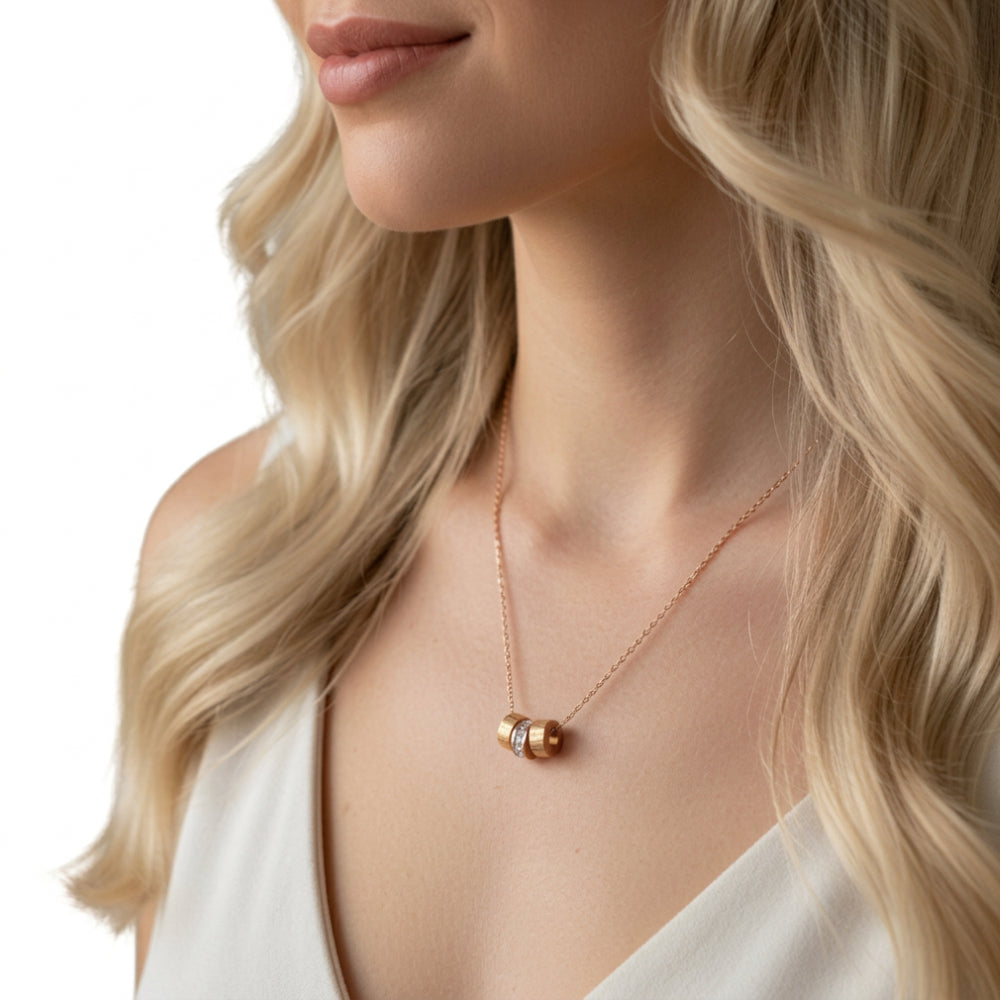 A close-up lifestyle shot of a woman wearing a delicate rose gold necklace with a small cylindrical bead pendant, styled with a white top against a clean white background.