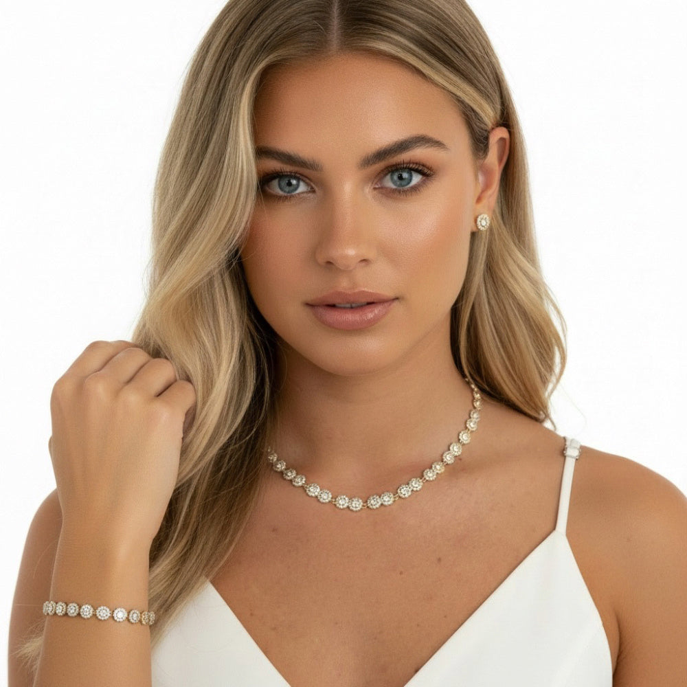 A blonde woman wearing a delicate gold-toned necklace and matching bracelet with clear stones, paired with a white camisole against a clean white background.