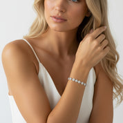 A close-up lifestyle shot of a model wearing a delicate silver bracelet adorned with small, round clear stones, paired with a white camisole against a clean white background.