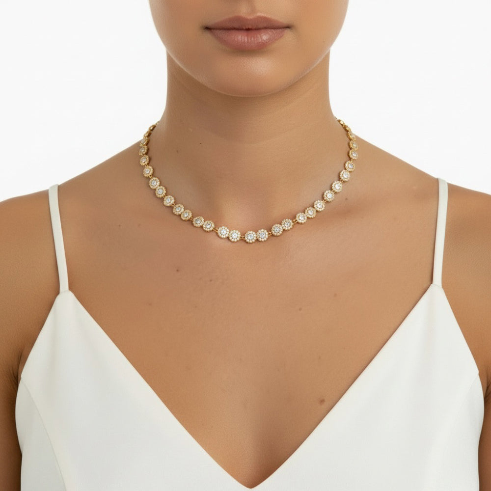 A close-up of a model wearing a delicate gold chain necklace adorned with small, round, clear pavé-set stones, styled with a white spaghetti-strap top against a clean white background.