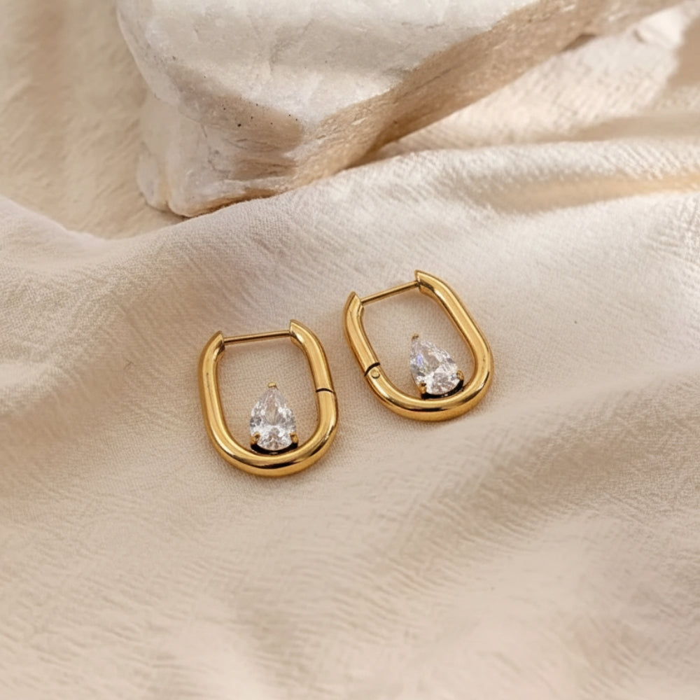 A pair of gold huggie hoop earrings with a small clear teardrop crystal, displayed on a textured beige fabric background.