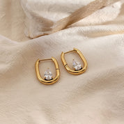 A pair of gold huggie hoop earrings with a small clear teardrop crystal, displayed on a textured beige fabric background.