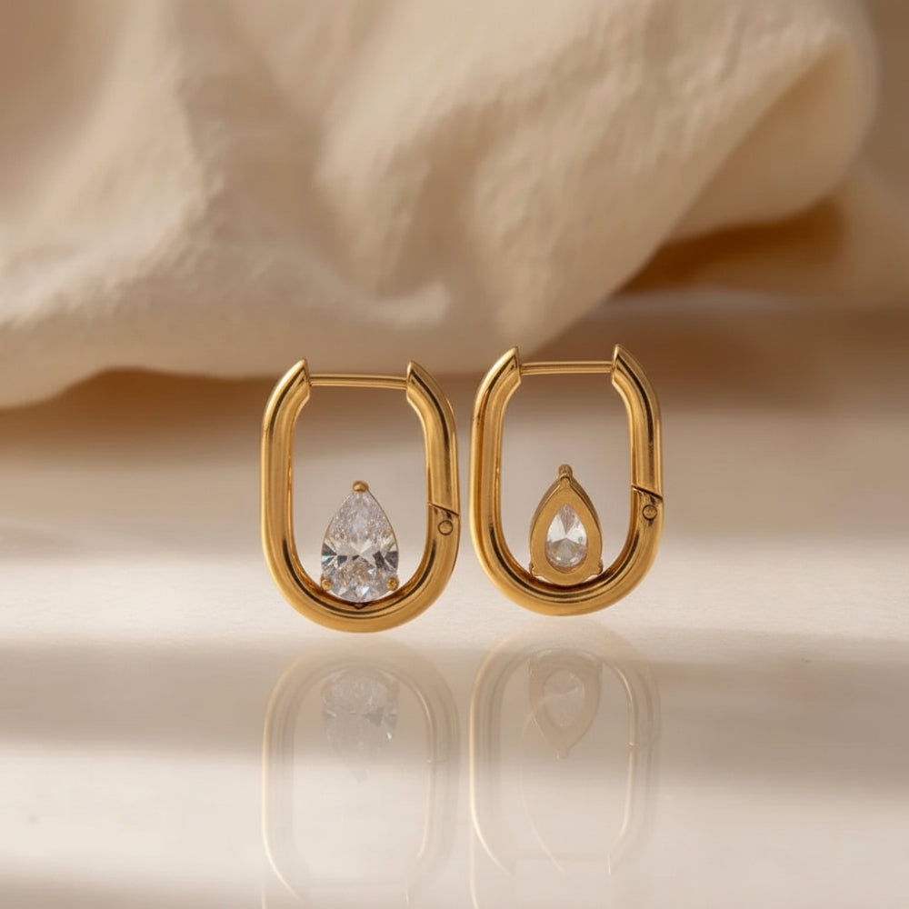 A pair of gold hoop-style earrings with teardrop-shaped clear gemstones, displayed on a reflective surface with a soft beige fabric background.