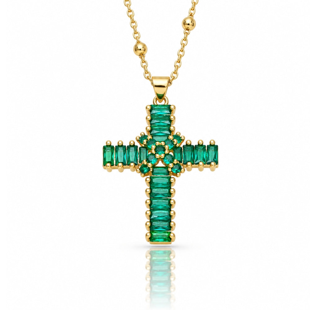 A gold cross pendant necklace featuring a row of emerald-cut green stones, displayed on a clean white background with a soft reflection below.