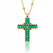 A gold cross pendant necklace featuring a row of emerald-cut green stones, displayed on a clean white background with a soft reflection below.