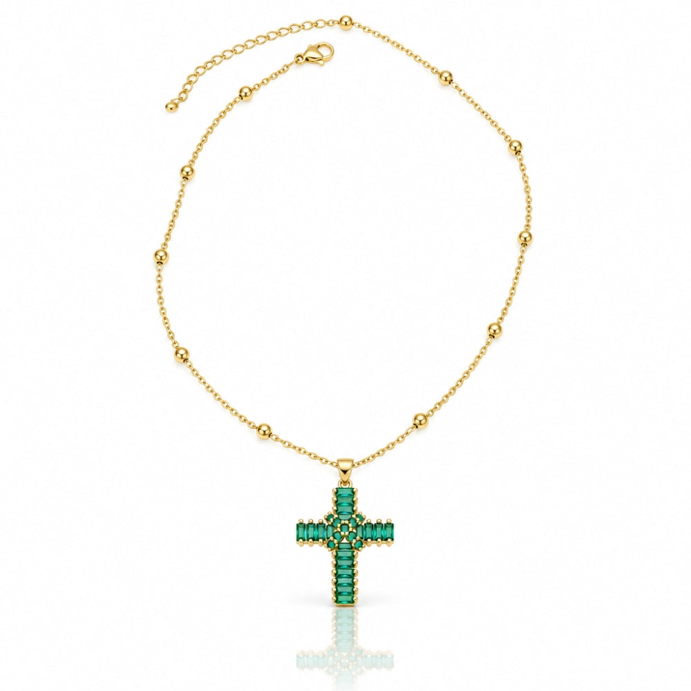 A gold chain necklace with a cross pendant adorned with green gemstones, displayed on a clean white background with a soft reflection below.