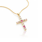 A gold cross pendant necklace adorned with pink and clear rectangular gemstones, suspended from a gold chain on a white background with a soft reflection.