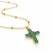 A gold cross pendant necklace adorned with baguette-cut green emerald stones, displayed on a clean white reflective surface.