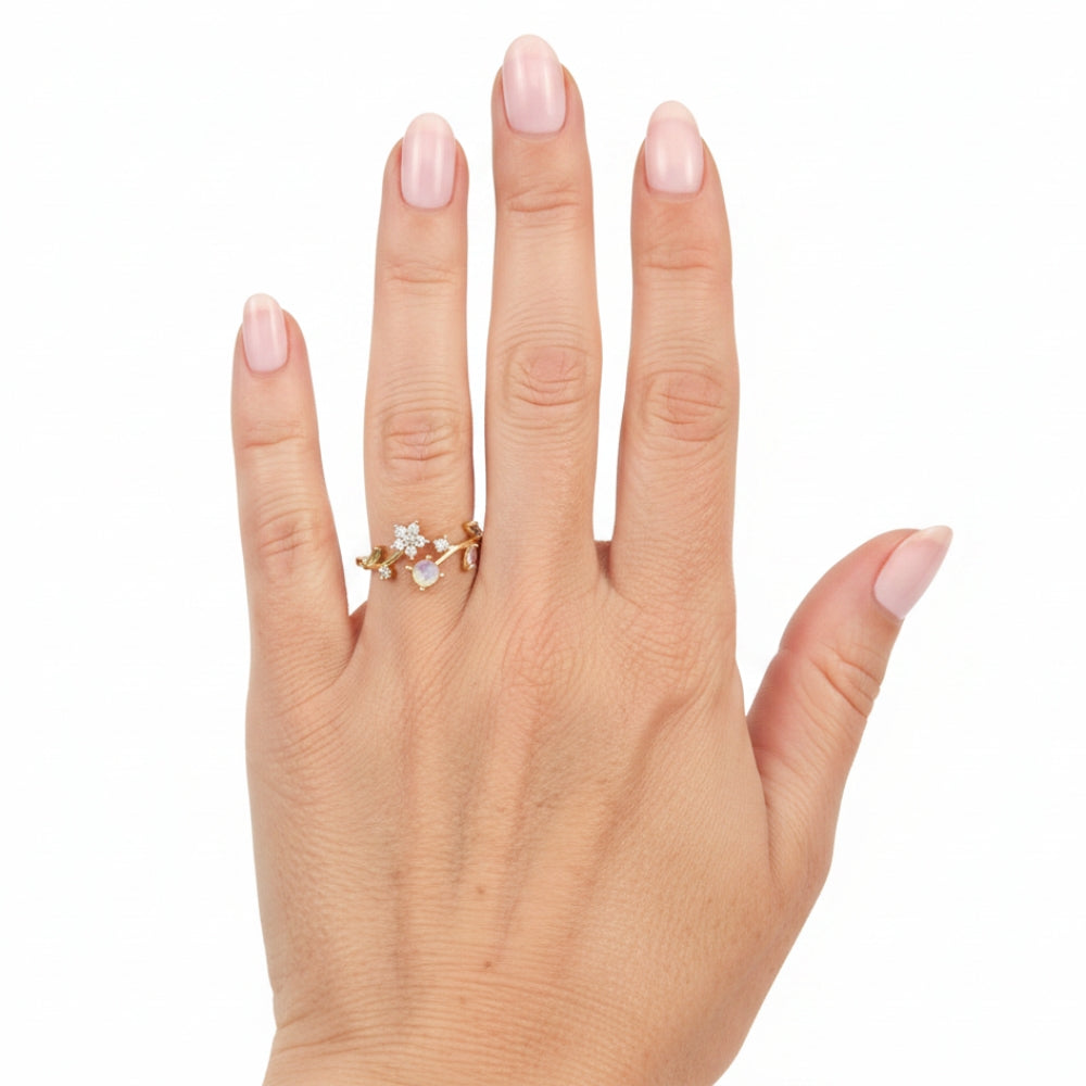 A close-up of a hand with pale pink manicured nails wearing a delicate gold ring with small clear stones, set against a plain white background.