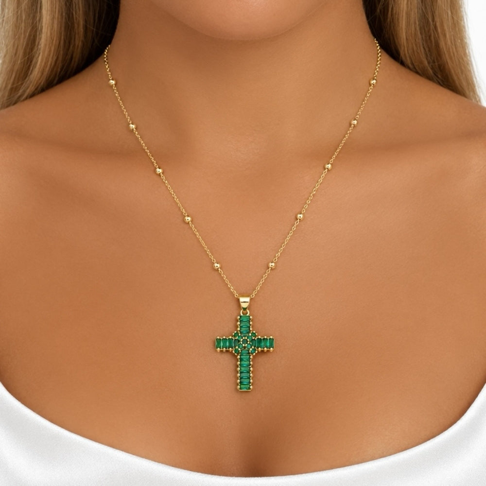A gold cross pendant necklace with green gemstone detailing, worn on a model's neck over a white top.
